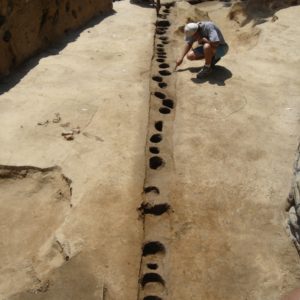 two archaeologists point to features in a long line of excavated post molds