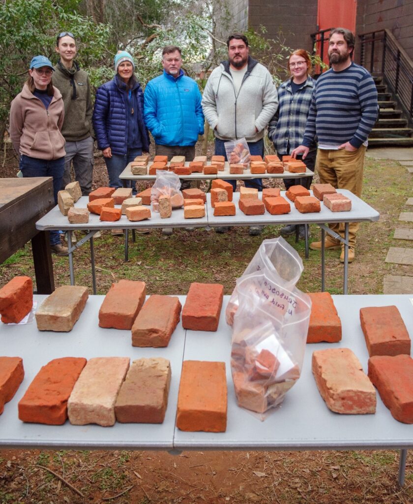 Seven people in cold-weather gear stand behind three tables. On the tables are many bricks, laid out individually for examination. Some brick fragments in plastic bags are nearby.