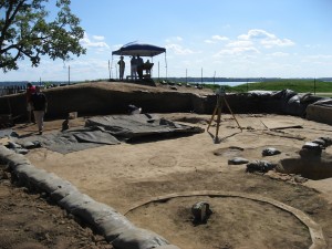 Archaeologists walking around the perimeter of an excavation unit containing a transit and various features