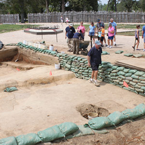 a group of visitors watch excavators screening artifacts and working in a large area lined with sandbags