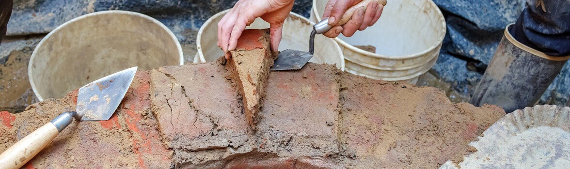 An archaeologist removes a wedge-shaped brick from the brick well ring using their hand and a trowel. The brick is covered in the original mortar.
