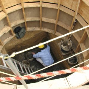 Archaeologist excavating a square well surrounded by a circular supporting wooden frame