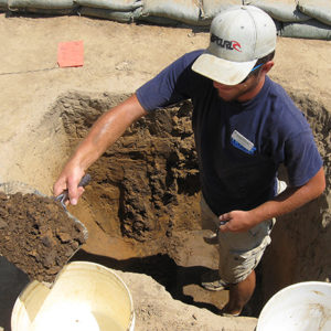 archaeologist standing in an excavation unit and pouring a dustpan of soil into a bucket