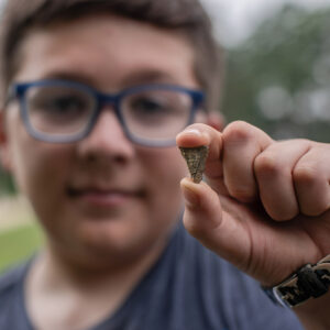 A camper holds the piece of eight that he found while screening. He is showing the reverse side of the coin. Some of the crown is visible.