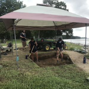 Archaeologists excavating with shovels under a small tent next to a riverbank