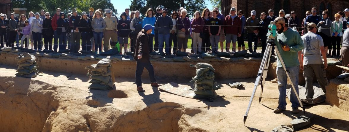 Large group listens to tour guide while archaeologists record finds
