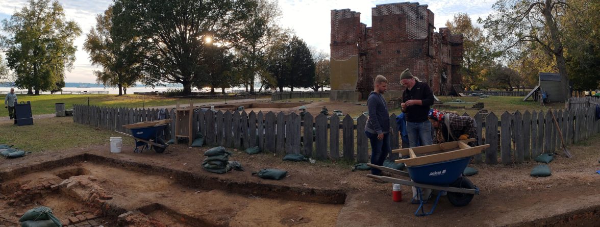 Archaeologists examine artifact while standing in excavations in front of brick ruins