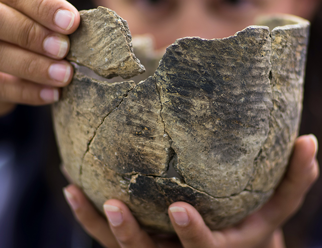 Hands holding mended sherds of a round-bottomed clay pot with stamped exterior